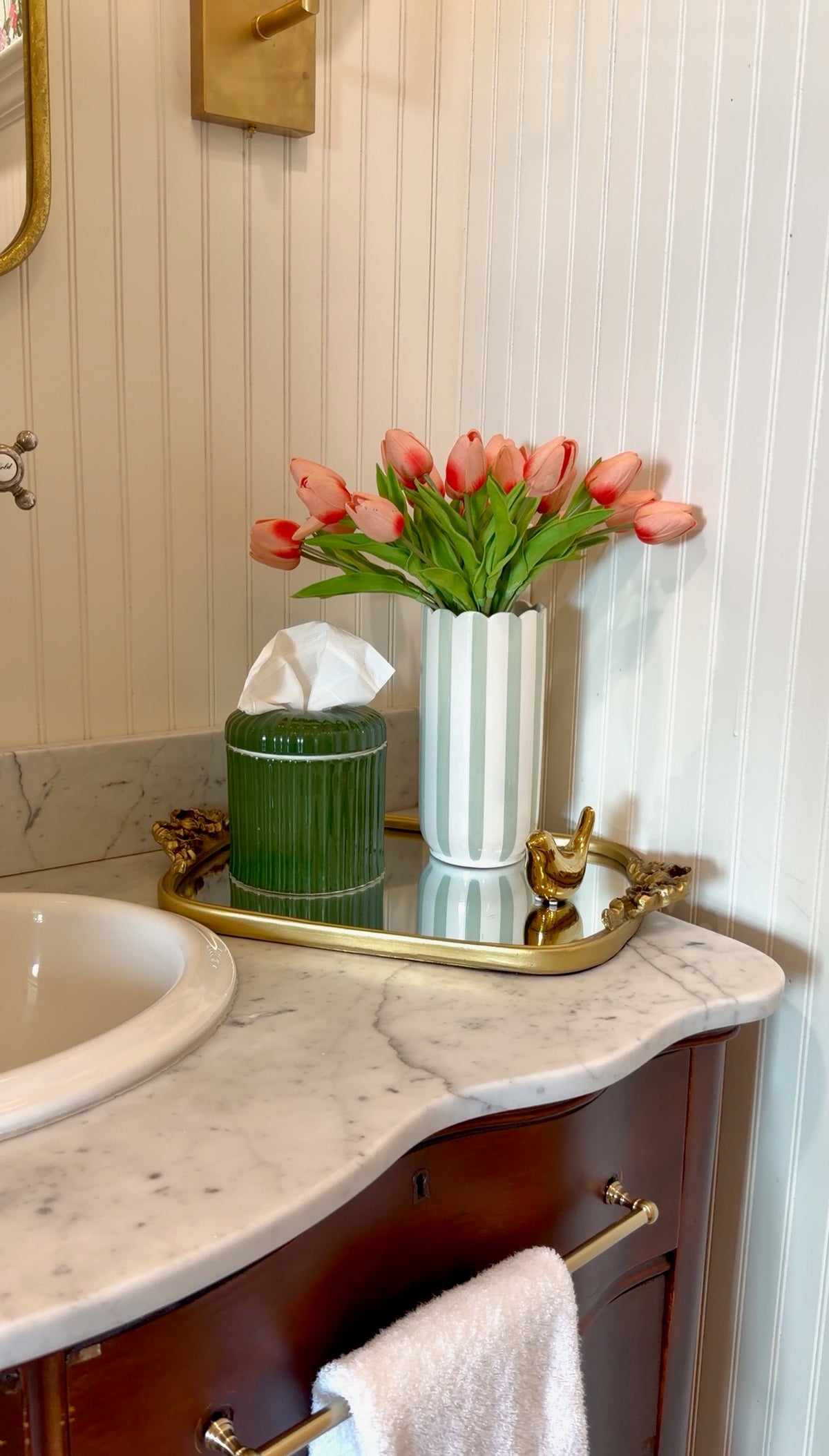 A styled bathroom vanity scene with a white marble countertop, dark wood cabinet, gold hardware, a mirror, a tissue box, a green ribbed container, and a white striped vase filled with pink tulips against a white paneled wall.