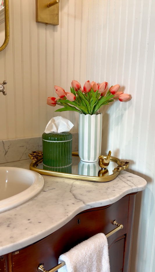 A styled bathroom vanity scene with a white marble countertop, dark wood cabinet, gold hardware, a mirror, a tissue box, a green ribbed container, and a white striped vase filled with pink tulips against a white paneled wall.