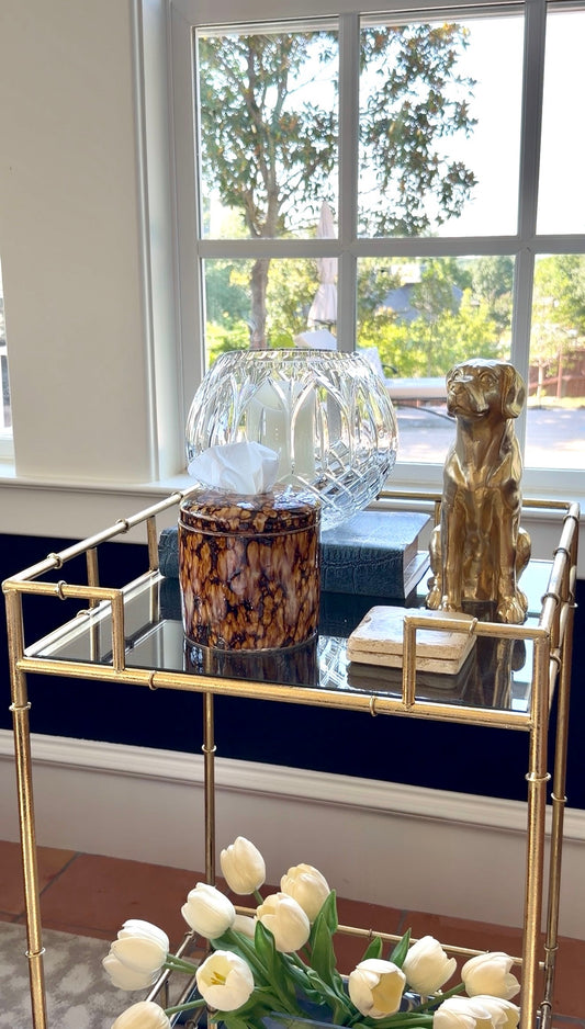 A gold-framed glass-top cart styled with home decor items, including a clear faceted glass bowl, a brown patterned tissue box cover, a blue book, a gold dog figurine, and a stack of books, set in front of a window with white tulips in the foreground.